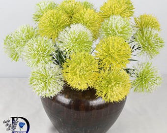 May include: A bouquet of artificial allium flowers in shades of lime green and white, arranged in a dark brown, rounded ceramic vase. The spherical flower heads feature thin, radiating petals, and the vase has a smooth, glossy finish.
