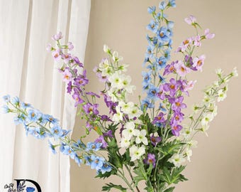 May include: A bouquet of artificial delphinium flowers featuring tall stems with clusters of small, five-petaled blossoms in shades of light blue, lavender, purple, and white, accented by green leaves and thin, branching stalks.