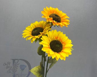 May include: Three vibrant sunflowers with bright yellow petals and dark brown centers. The artificial flowers have green stems and leaves, set against a gray backdrop. The flowers are in full bloom, showcasing their natural beauty.