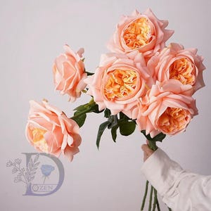 May include: A bunch of peach-coloured roses with vibrant orange centres. The roses are in full bloom, with multiple petals creating a layered effect. Green stems and leaves provide contrast against the soft, light background.