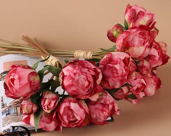 May include: A bouquet of artificial peony flowers featuring multiple shades of pink and red petals with green stems. The flowers are bundled together with a light-colored raffia tie. The stems are resting on an open book with printed text.