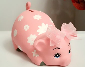 Könnte beinhalten: Rosa Keramik-Sparschwein mit weißen Blumendesigns und einem rosa Band. Das Sparschwein hat blaue Augen und eine bemalte Schnauze.