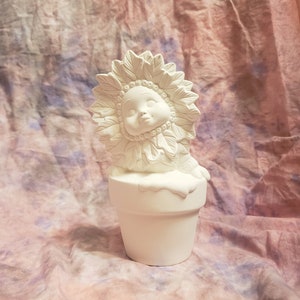 Puede incluir: Figurilla de cerámica blanca de un girasol con cara, que lleva una maceta como sombrero. La figura está en posición sentada.
