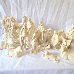 Puede incluir: Un conjunto de figuras de belén de cerámica blanquecina. El conjunto incluye figuras de personas, animales como un camello, un burro y una vaca, y otros elementos tradicionales. Las figuras están dispuestas sobre una superficie blanca.
