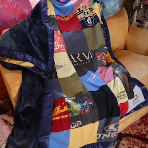 May include: A blue and navy blue patchwork quilt made from old t-shirts. The quilt is made up of various colors and patterns, including red, yellow, green, and white. The quilt is sitting on a couch with a pillow on the back of the couch.