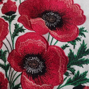 Puede incluir: Amapolas rojas bordadas con hojas y tallos verdes sobre un fondo de tela blanca.