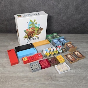 Puede incluir: Un juego de mesa "EMBERLEAF" con una caja que presenta una ilustración de un ratón, un molino de viento y un paisaje. El juego incluye varias bandejas de colores, cartas y piezas de juego, dispuestas sobre una superficie de madera.