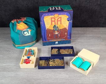 May include: A board game called "Ra" with a blue, yellow, and red box featuring an illustration of the Egyptian god Ra. The game includes a teal drawstring bag, a cream-colored tray with compartments, and two blue trays with compartments filled with gold-colored tokens.