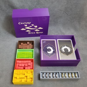 Puede incluir: Caja púrpura con una tapa que dice "Century Spice Road" y contiene compartimentos para guardar piezas de juego. La caja tiene cuatro compartimentos para cubos en marrón, verde, rojo y amarillo. Hay un compartimento para cartas y un compartimento para monedas.