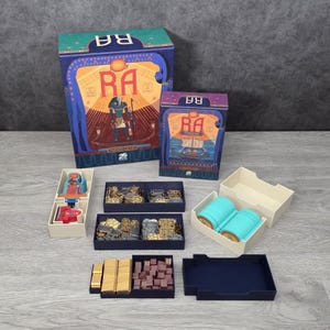 Puede incluir: Juego de mesa titulado "Ra" con una caja que presenta una ilustración de una figura egipcia. El juego incluye varias piezas en bandejas, incluyendo fichas doradas, cilindros azules y componentes rojos y beige. La caja es azul, verde y morada.