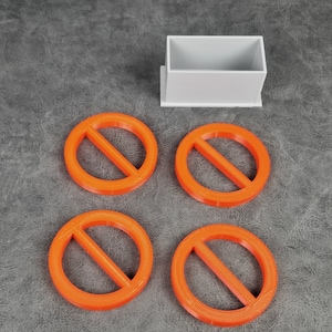 Può includere: Quattro oggetti circolari arancioni brillanti con una linea diagonale che li attraversa, simili a un simbolo di "vietato", sono disposti vicino a un piccolo contenitore rettangolare grigio chiaro. Gli oggetti sono probabilmente stampati in 3D e hanno una superficie strutturata.