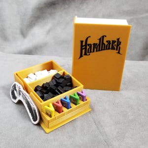以下が含まれることがあります： ゲームの駒やカードを入れるための木製オーガナイザー。オーガナイザーには「ハードバック」とラベルが貼られており、側面には白黒の羽根のデザインが施されています。