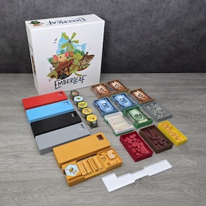 Puede incluir: Juego de mesa Emberleaf con una caja blanca con una ilustración de un ratón y un molino de viento. Incluye bandejas de colores, fichas hexagonales y cartas de juego. Los componentes del juego están dispuestos sobre una superficie gris.