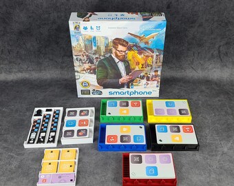 May include: A board game called "Smartphone" with a colorful box and game pieces. The box features a cartoon image of a man in a suit using a smartphone. The game pieces are made of plastic and have different colors and shapes.