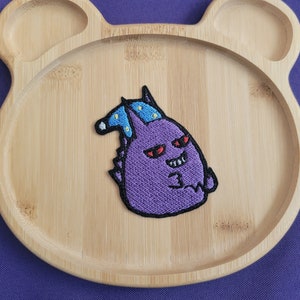 Peut inclure: Patch brodé violet d'un personnage de dessin animé portant un chapeau bleu avec des étoiles jaunes. Le personnage a des yeux rouges et un sourire malicieux.