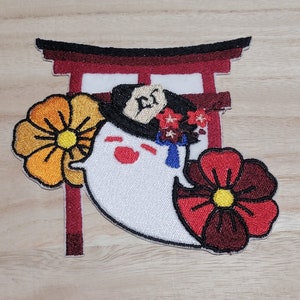 Puede incluir: Parche bordado con un fantasma blanco que lleva un sombrero negro con una flor roja y una cinta azul. El fantasma está de pie frente a una puerta torii roja con una flor amarilla a la izquierda y una flor roja a la derecha.