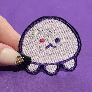 May include: A purple embroidered patch of a cartoon ghost with a sad face. The ghost has two black eyes, a pink nose, and four small legs.
