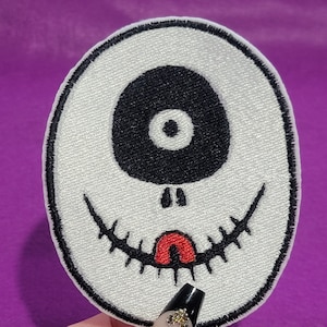 Könnte beinhalten: Weißer, ovaler gestickter Aufnäher mit einem schwarzen Auge und einem roten Mund. Das Auge hat eine weiße Pupille. Der Mund ist mit einer gezackten Linie gestickt und hat eine rote Zunge.