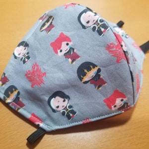 Puede incluir: Una mascarilla facial de tela gris con un patrón repetido de personajes de dibujos animados y diseños de dragones rojos. La mascarilla tiene lazos elásticos negros para las orejas.