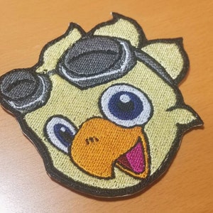 May include: Embroidered patch of a yellow bird wearing goggles, with a blue eye and orange beak. The bird is smiling with a pink tongue.