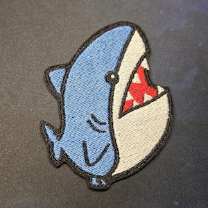 May include: Embroidered patch featuring a cartoon shark with a wide open mouth. The shark is blue and white with a red mouth and black outline. The patch is likely for clothing or accessories.