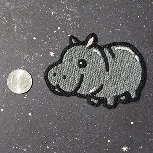 May include: A gray embroidered iron-on patch of a cartoon hippopotamus with a pink nose and a white eye.