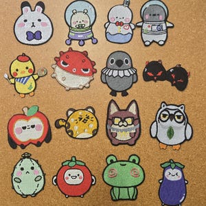 May include: A collection of sixteen embroidered patches featuring various cartoon characters, including animals, fruits, and vegetables. The designs include a rabbit, astronaut, pufferfish, bird, apple, leopard, owl, cactus, tomato, frog, and eggplant.