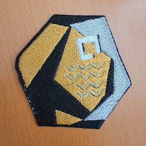 May include: A black, gold, and silver embroidered patch with a geometric design. The patch features a stylized shape with wavy lines and a square in the center.