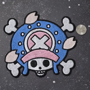 May include: A blue and white embroidered patch with a skull and crossbones, a pink hat, and a white X in the center. The patch is surrounded by a circle of white dots.