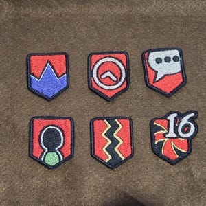 May include: Six embroidered patches with red, black, and white designs. The patches feature a crown, a chevron, a speech bubble, a stick figure, a zig-zag pattern, and a number 16.