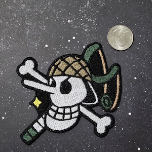 May include: White skull and crossbones with a green helmet and a green and brown sword. The skull is smiling and has a single eye. The background is a dark gray with white speckles.