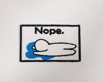 May include: White embroidered patch with a black outline. The patch features a cartoon character lying in a blue puddle with the text "Nope."