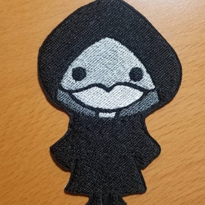 Puede incluir: Parche bordado en blanco y negro de una figura encapuchada con una cara pequeña y redonda y ojos grandes. La figura lleva una larga túnica con capucha.
