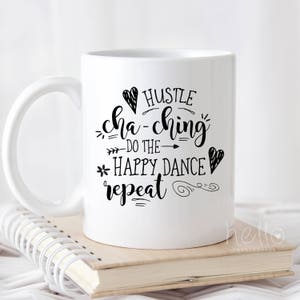 K&ouml;nnte beinhalten: Wei&szlig;e Keramik-Kaffeetasse mit schwarzem Text, der "Hustle cha-ching -do the happy dance- repeat" lautet.
