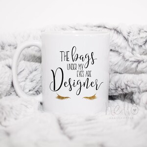May include: White ceramic coffee mug with a black and gold design. The mug reads "The bags under my eyes are designer" with a pair of gold eyelashes below the text.