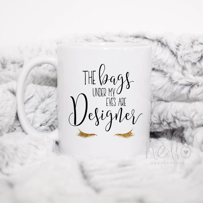 May include: White ceramic coffee mug with a black and gold design. The mug reads "The bags under my eyes are designer" with a pair of gold eyelashes below the text.