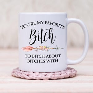 May include: White ceramic mug with the text "YOU'RE MY FAVORITE Bitch TO BITCH ABOUT BITCHES WITH" in black script. The design includes a floral arrow. The mug sits on a pink crocheted coaster.