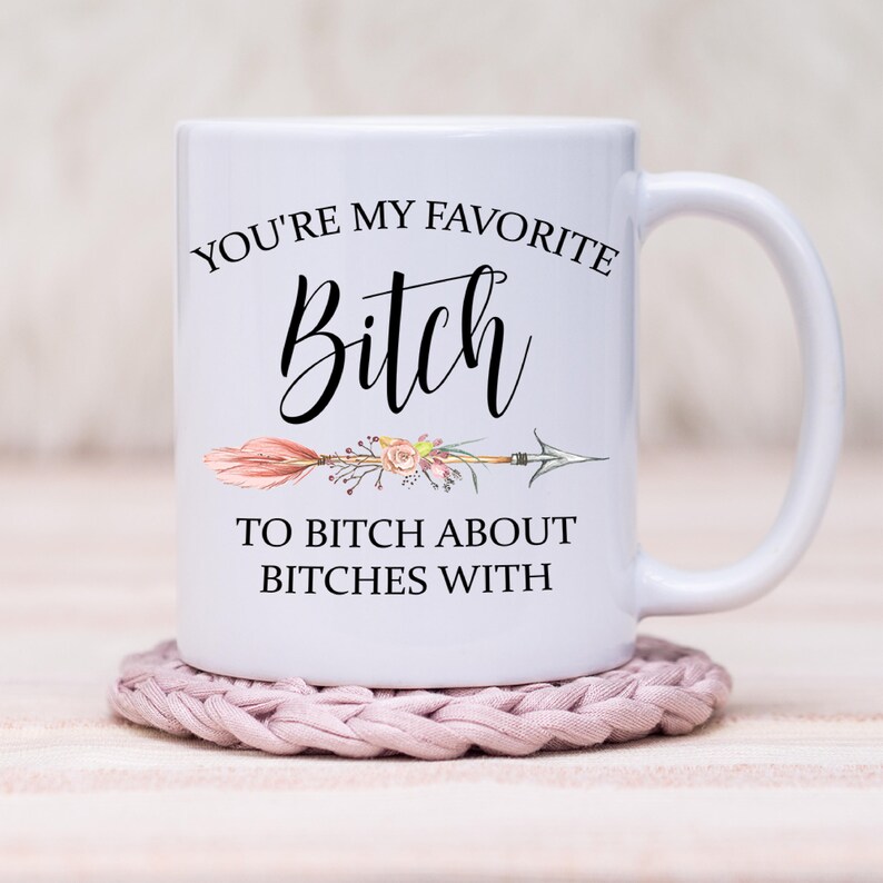 May include: White ceramic mug with the text "YOU'RE MY FAVORITE Bitch TO BITCH ABOUT BITCHES WITH" in black script. The design includes a floral arrow. The mug sits on a pink crocheted coaster.