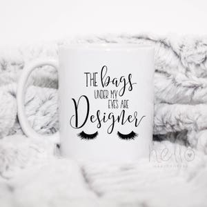 May include: White ceramic mug with black text that reads "The bags under my eyes are designer" with two black eyelashes below the text.