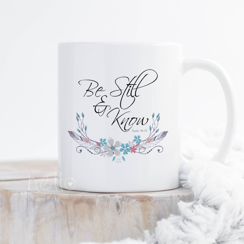 May include: White ceramic mug featuring the words "Be Still & Know" in a stylish script, complemented by a floral and feather design in blue, pink, and grey. The mug has a curved handle and is suitable for hot or cold drinks.