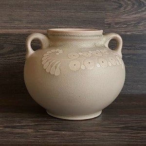 May include: A beige ceramic vase with two handles and a floral design. The vase has a textured surface and a slightly rounded shape.