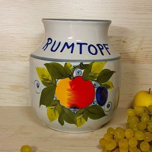 May include: White ceramic jar with a blue and green painted design of fruit and leaves. The jar has the word "RUMTOPF" written on it in blue letters. The jar is sitting on a wooden surface with a bunch of green grapes and a yellow apple.