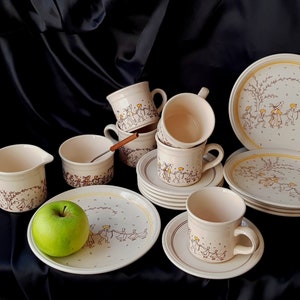May include: A vintage set of ceramic teacups and saucers featuring a charming design of children and geese. The cups and saucers are cream coloured with brown and yellow accents.