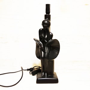 May include: A black ceramic table lamp shaped like a woman in a flowing dress. The lamp has a black cord and plug.
