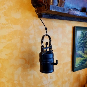 Könnte beinhalten: Eine schwarze Öllampe aus Metall, die an einem Haken an einer gelben Wand hängt. Die Lampe hat eine Kette und einen runden, zylindrischen Körper.