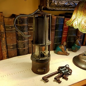 Könnte beinhalten: Eine antike Messing-Bergmannslampe mit einer Glaslinse und einem schwarzen Metallkäfig. Die Lampe hat einen Griff und steht auf einer weißen Oberfläche mit zwei alten Schlüsseln und einem Schlüsselanhänger mit der Nummer 32.