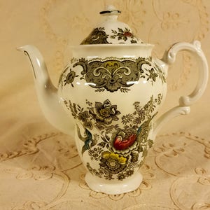 Pode incluir: Uma chaleira de cerâmica branca com um padrão floral em tons de verde, marrom, amarelo e vermelho. A chaleira tem uma tampa com uma borda dourada e uma alça.