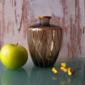 Op de afbeelding: Een donkergrijze glazen vaas met gouden accenten en een golvend patroon. De vaas staat op een houten oppervlak.