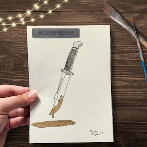 May include: A black and white drawing of a knife with a brown stain on the blade. The drawing is on a white background with the word "AICHMOPHOBIA" written in black at the top. The drawing is signed "Emily '23" in the bottom right corner.
