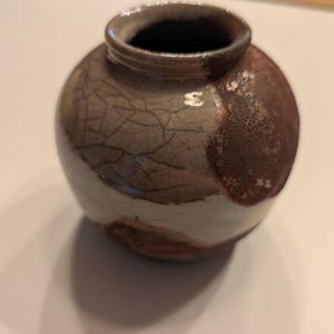 May include: A small, round ceramic vase with a wide opening and a short neck. The surface features a crackled glaze finish in shades of grey, white, and deep brown, with a textured, mottled appearance across the rounded body of the vessel.